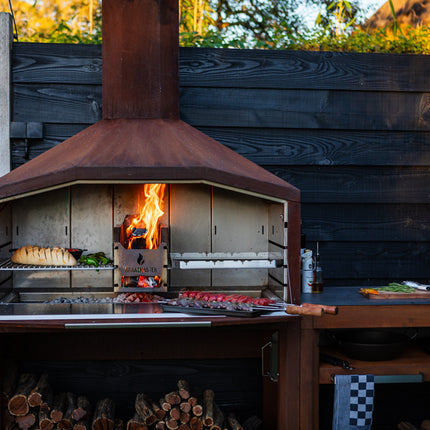 Braaimaster Duo Corten met zijtafel tegen een schutting met vuur in de firebox en brood op het rooster