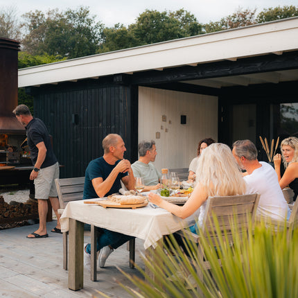 Braaimaster Duo Corten brandend op een terras met een braaimaster aan het werk en een gezellige diner tafel gevuld met mannen en vrouwen op de voorgrond