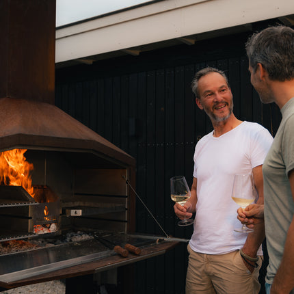 Braaimaster duo corten met vuur in de firebox en 2 mannen pratend op de voorgrond met beide een glas witte wijn