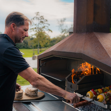 Braaimaster Duo Corten brandend in tuine met een braaimaster die churrasco spiesen draait