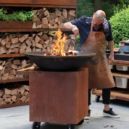 OFYR Heritage Classic 100 PRO Corten op terras met chef en brandend vuur en OFYR Woodstorage op de achtergrond