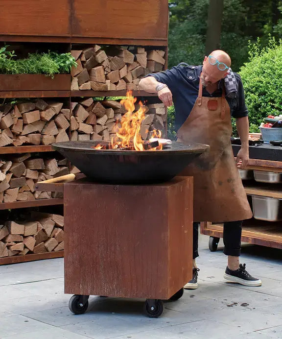 OFYR Heritage Classic 100 PRO Corten op terras met chef en brandend vuur en OFYR Woodstorage op de achtergrond