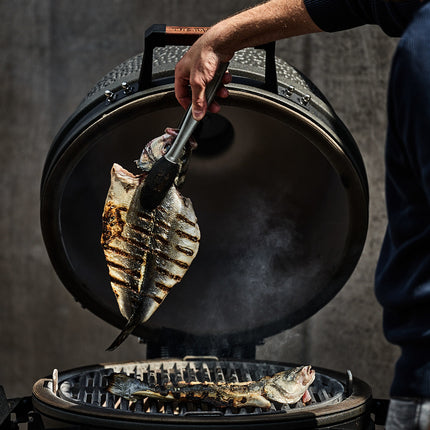 The Bastard kamado VX Large Solo met geopende deksel en een pitmaster die vis grilt op het gietijzeren rooster
